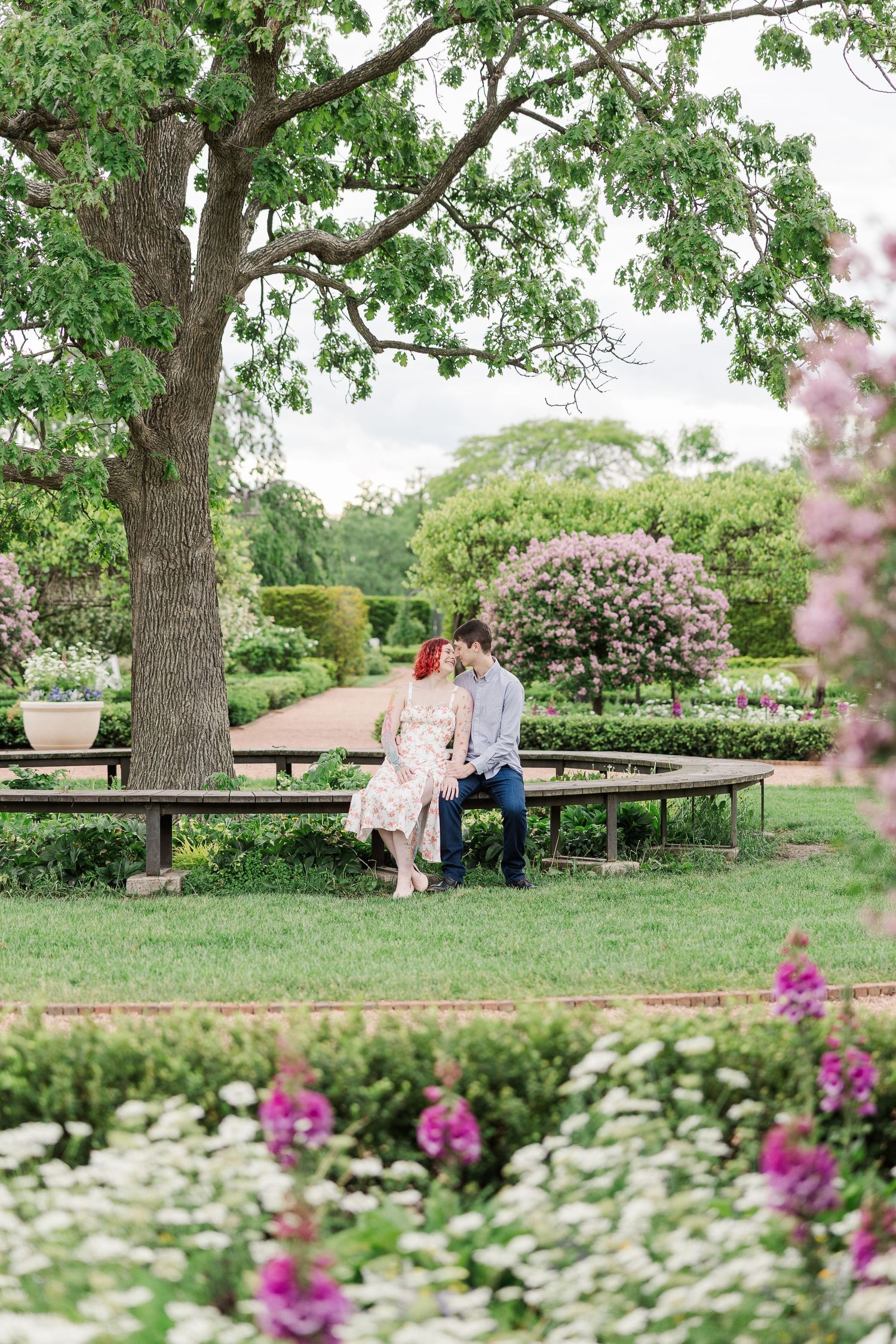 A couple sits together on a wooden bench in a lush garden with blooming pink and white flowers under a large tree at Cantigny Park