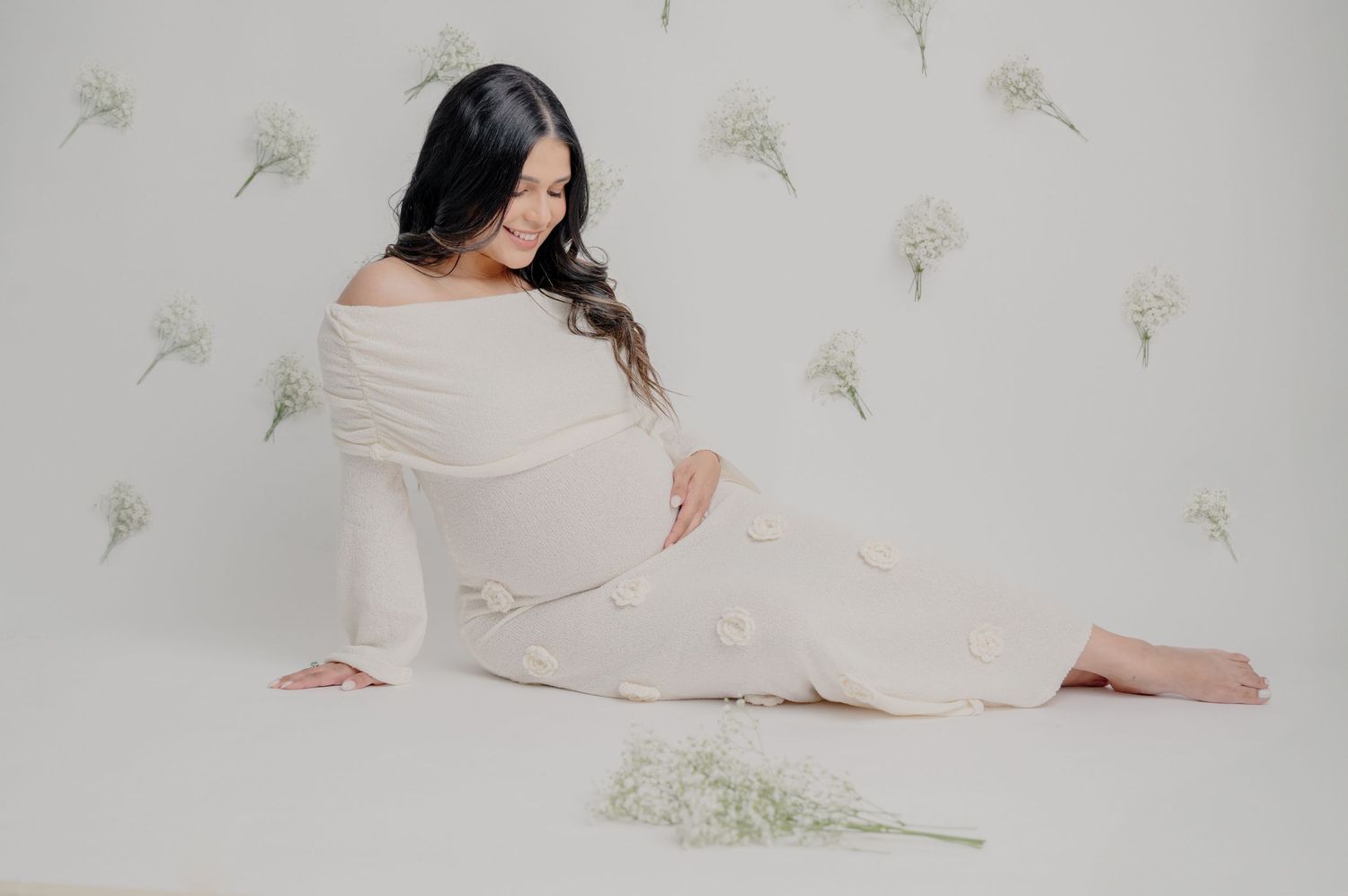 A person in a white flowing dress poses elegantly against a white background with delicate floral wall accents.