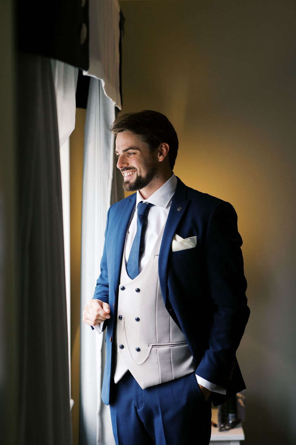 A groom in a navy blue suit and white vest stands near a window, creating a silhouetted profile shot in moody lighting.