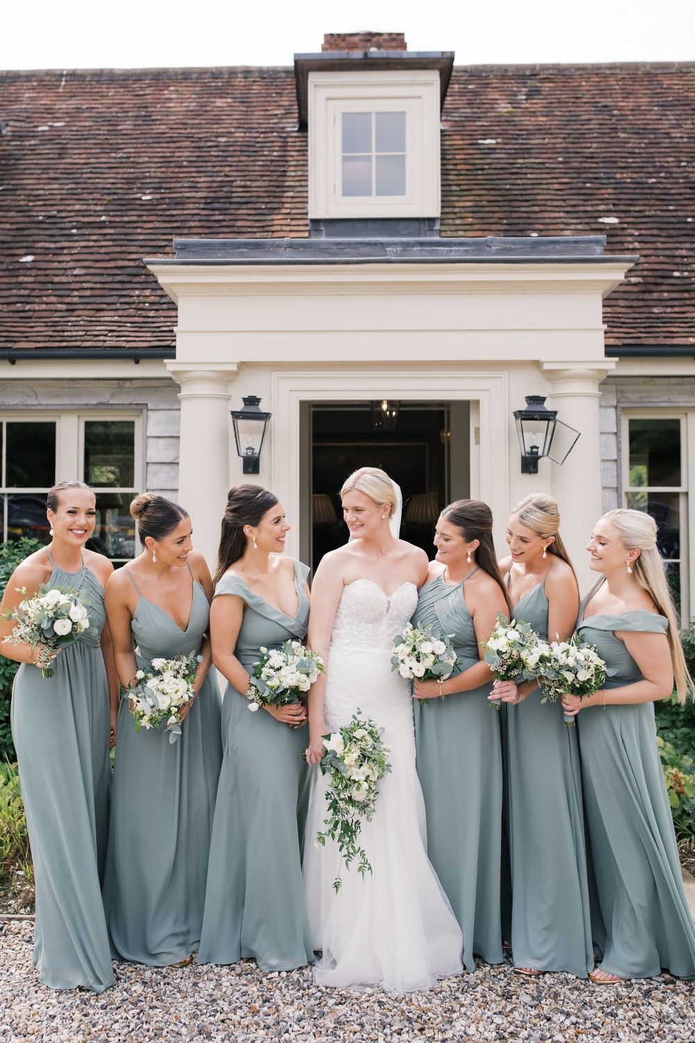 A bridal party in sage green dresses and white floral bouquets poses together outside a classic brick home.