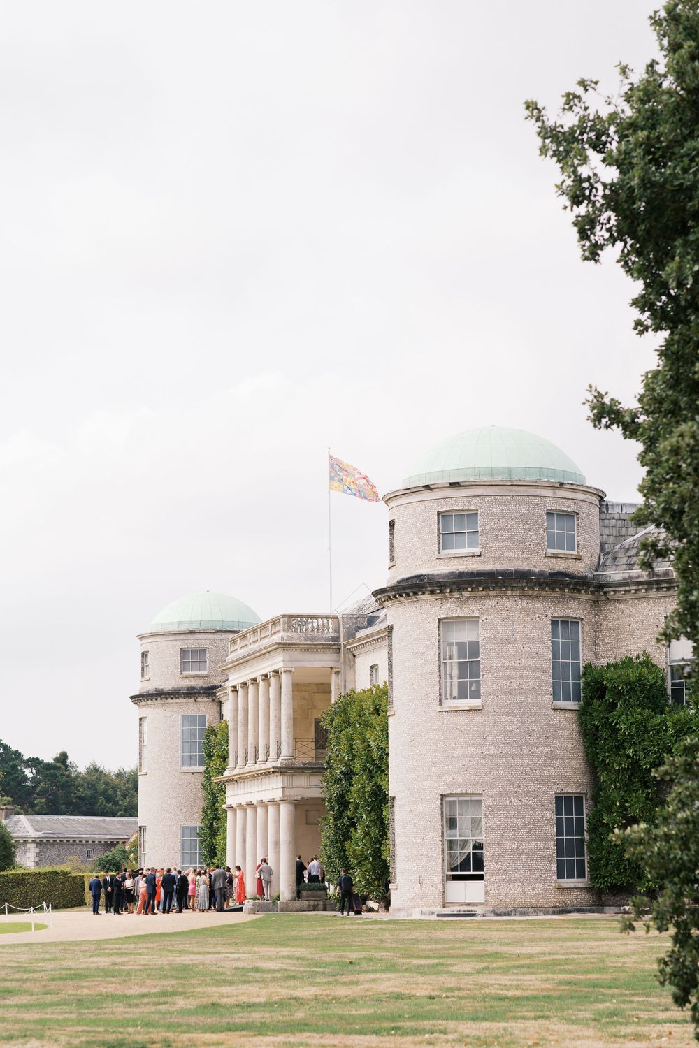 Panoramic view of a grand neoclassical mansion with curved wings and stone columns set against a cloudy sky.