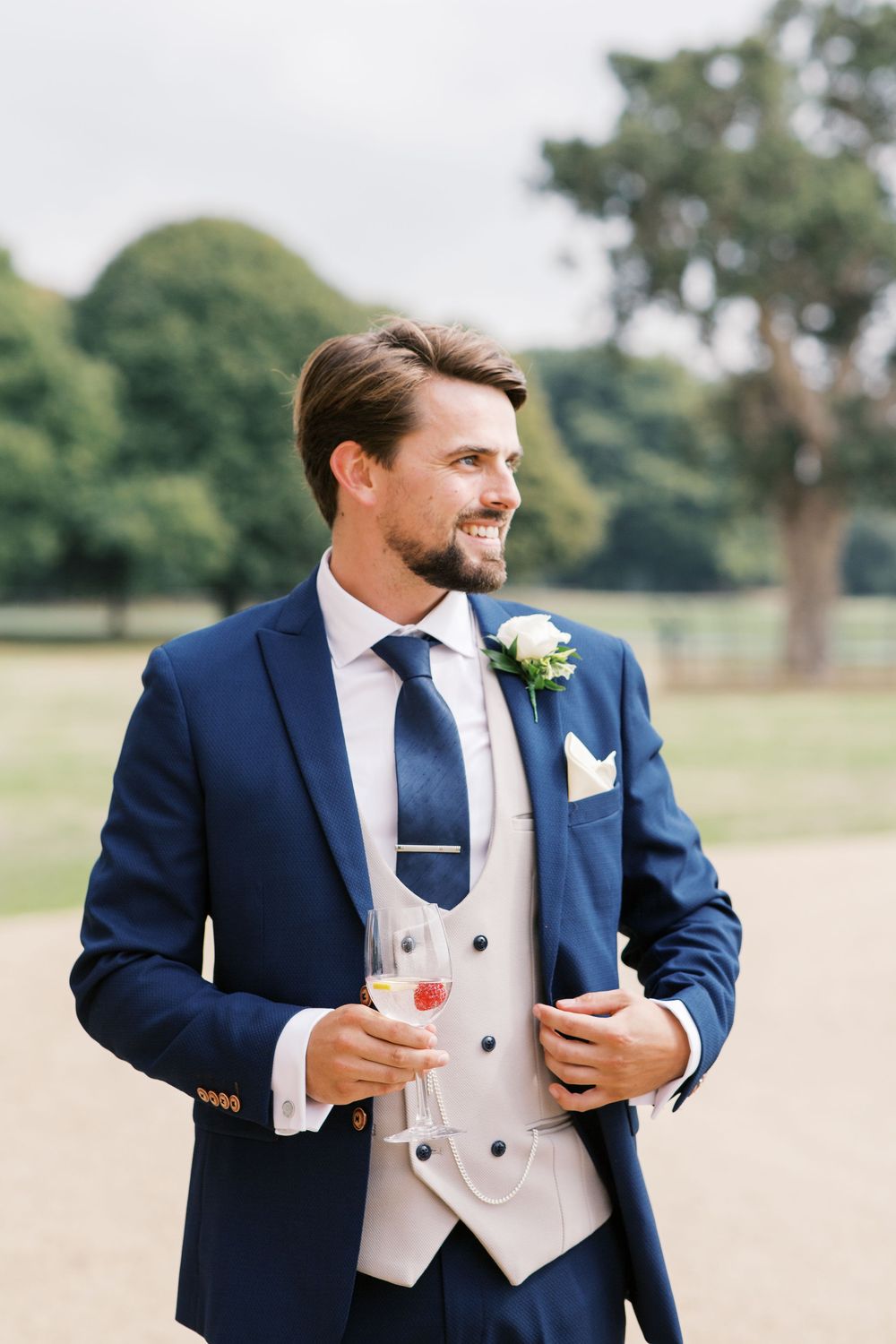 A groom wearing a navy blue suit with light gray waistcoat and white boutonniere smiles while standing outdoors.