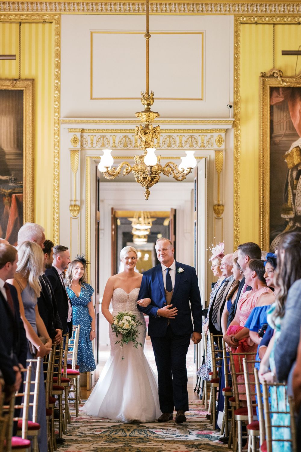 Elegant wedding ceremony in ornate ballroom with gold trim, chandeliers and guests lining the aisle.