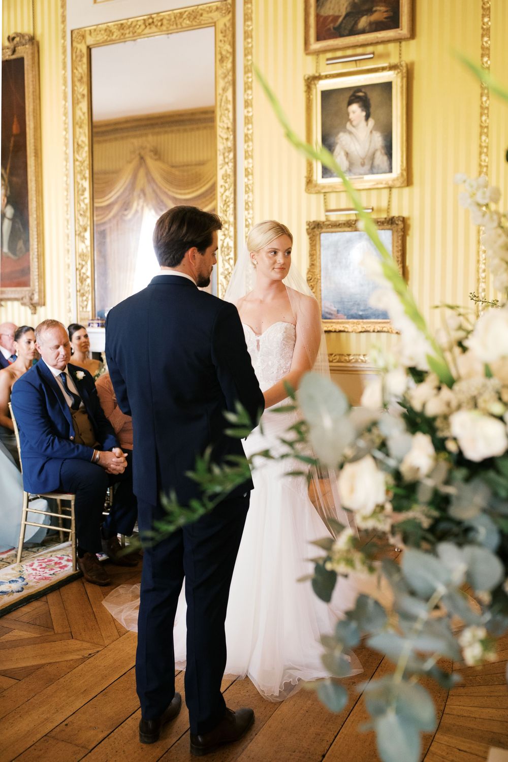 Elegant indoor wedding ceremony in a historic room with yellow walls, ornate gold frames, and white floral arrangements.