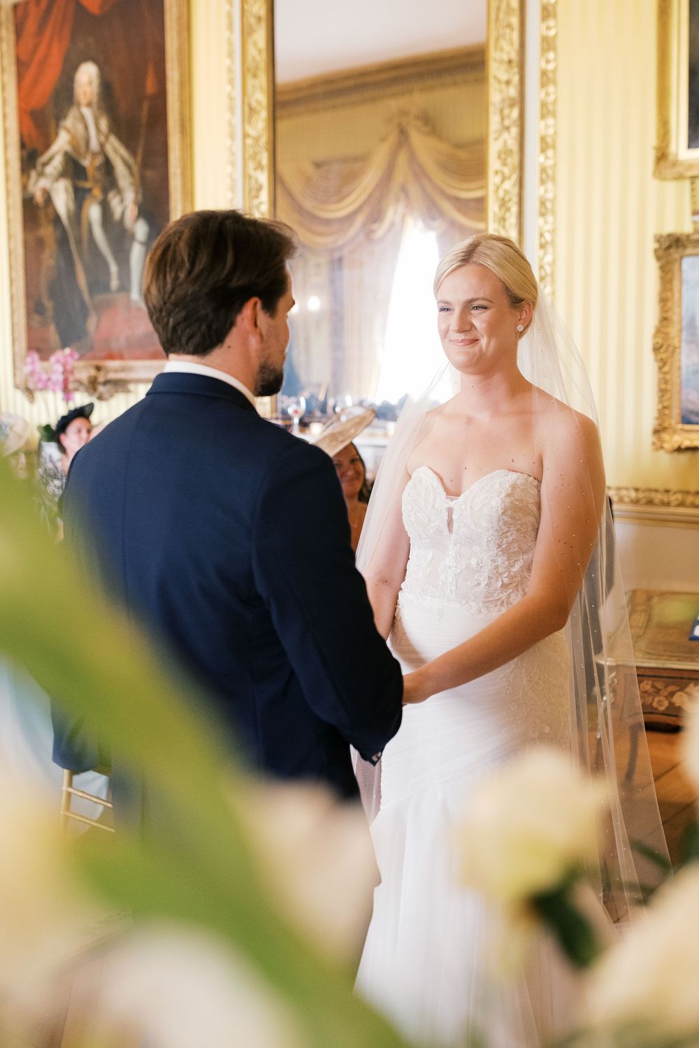 Elegant wedding ceremony takes place in an ornate room with portraits and floral arrangements.