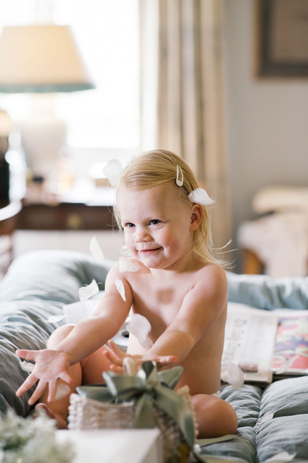 A toddler in natural light sits on a bed with gray bedding while playing with a cushion.