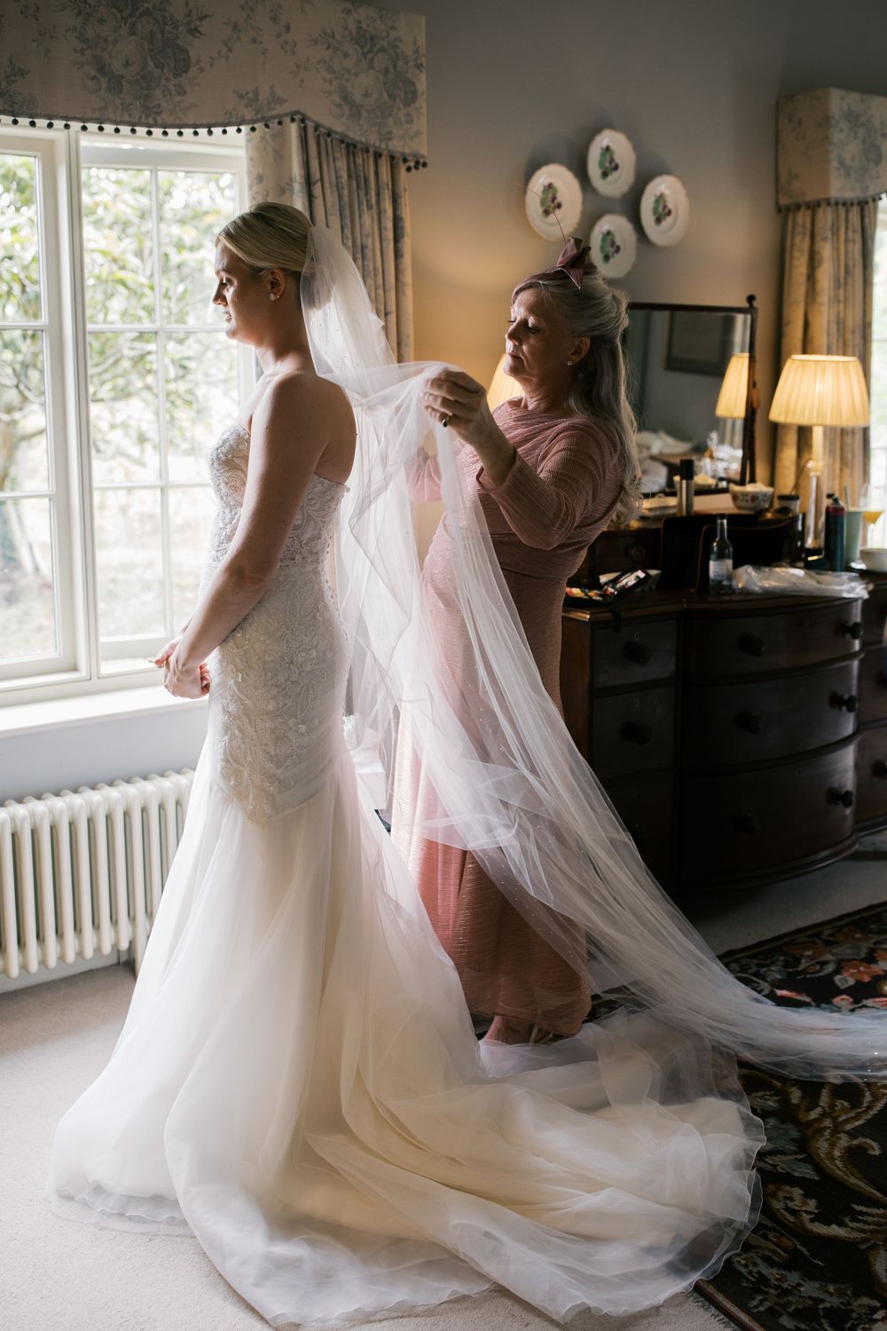 A long flowing white wedding gown and veil are being adjusted in an elegantly decorated room with a bay window.