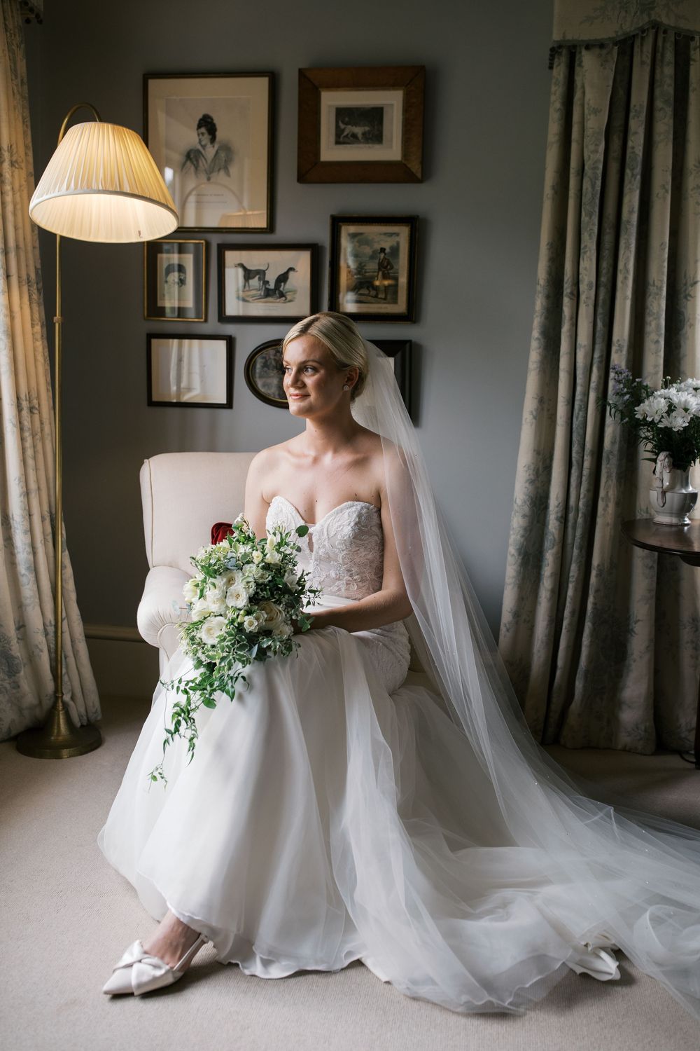 Elegant bridal portraits in soft lighting showcase a flowing white wedding gown and veil against a gray wall with framed artwork.