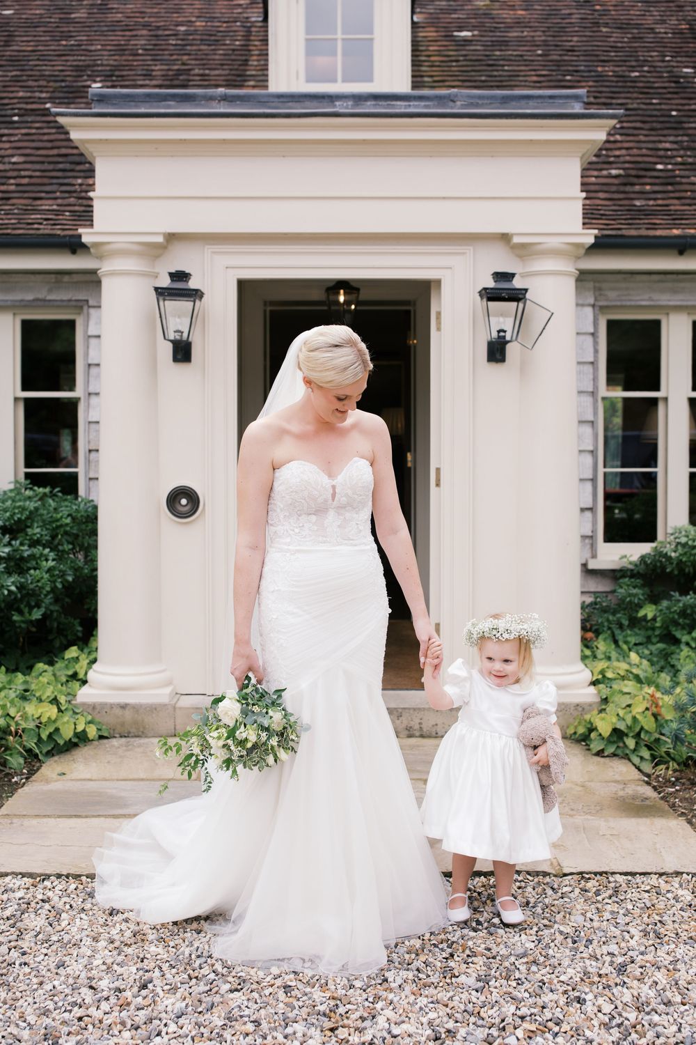 Classic wedding scene at an elegant white house with gravel driveway featuring bride and flower girl in matching white dresses.