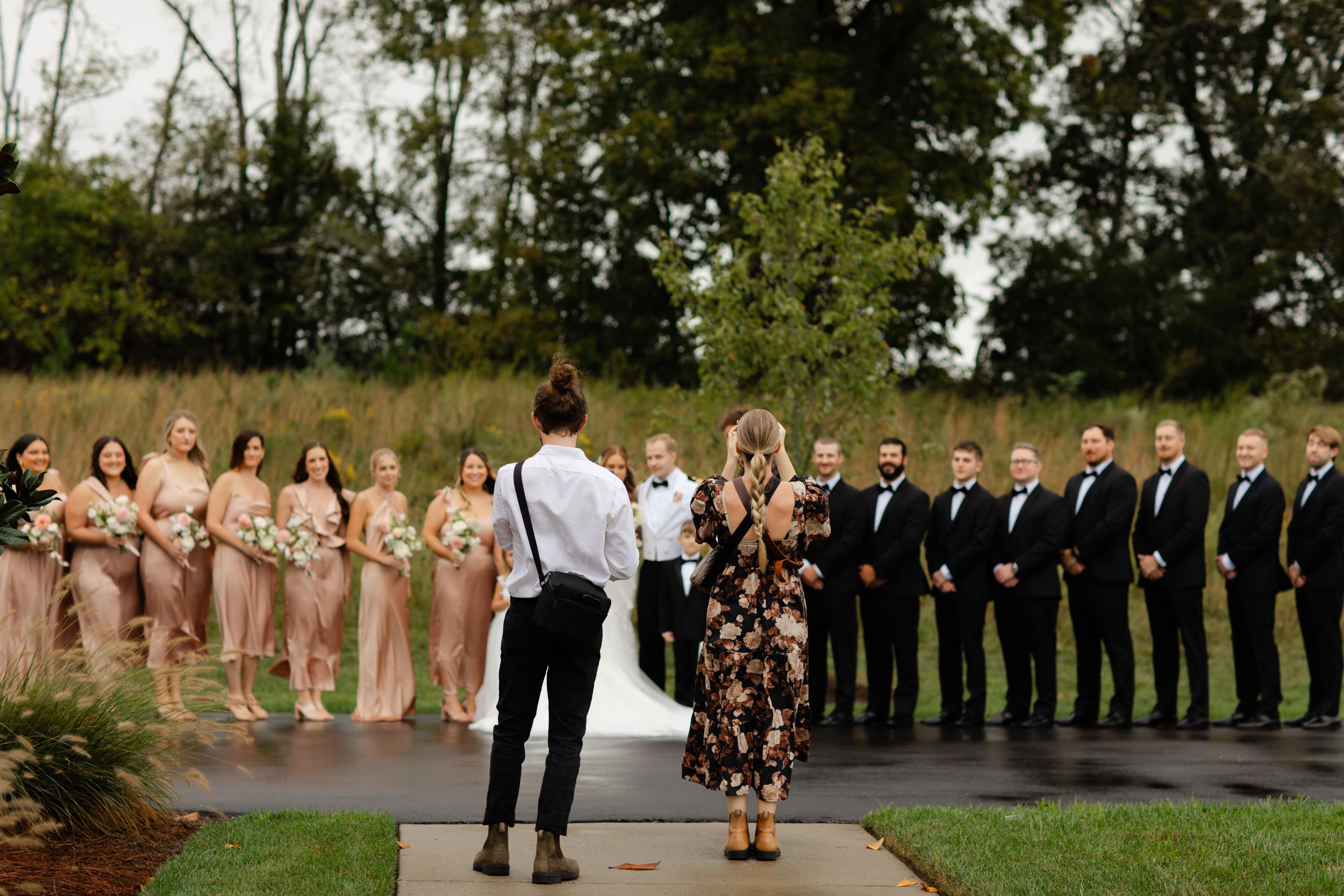 Wedding party in formal attire poses together outdoors on a sunny day with trees and greenery in the background.