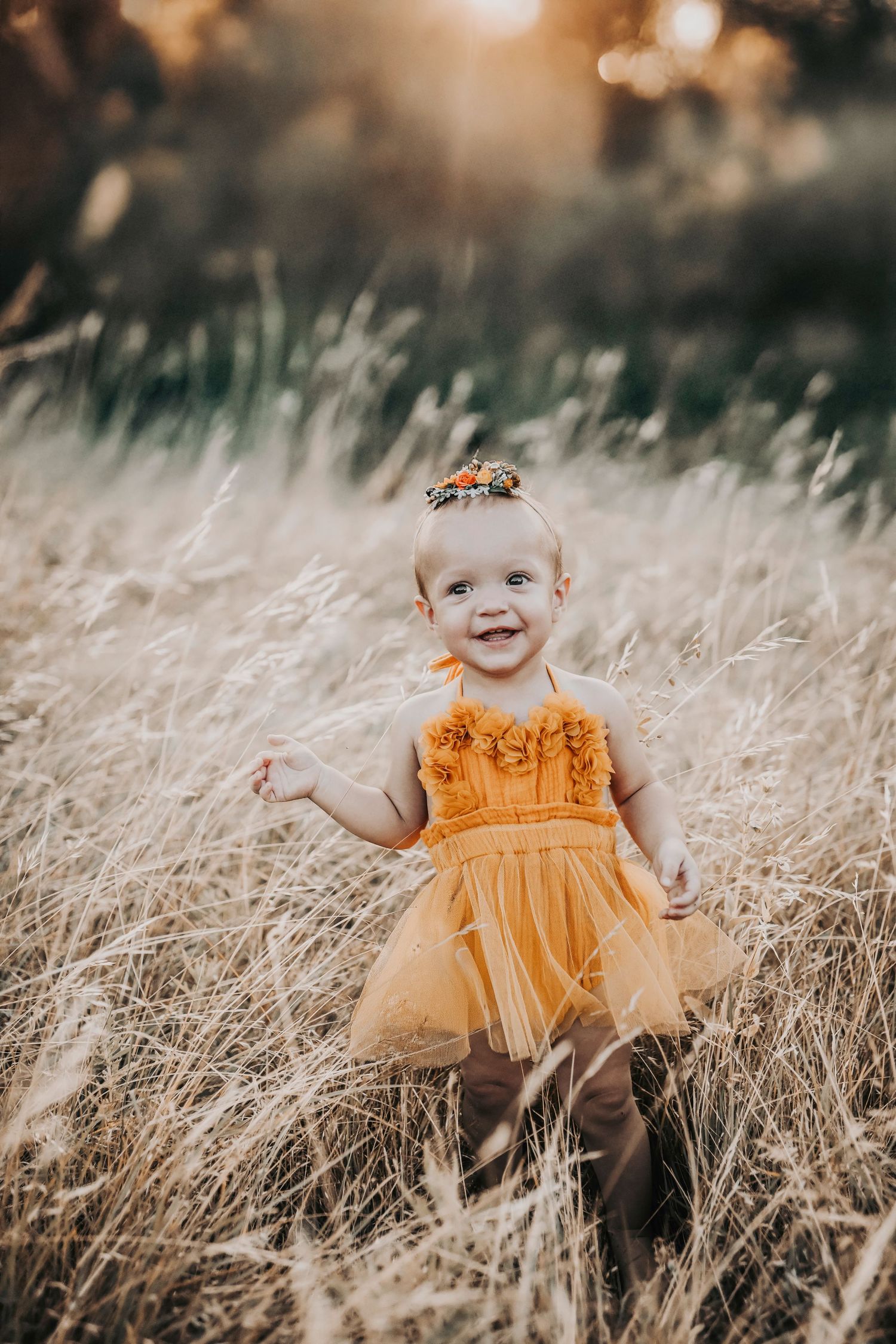 a very happy one year old girl in a burnt orange tutu dress smiles in a warm field at sunset