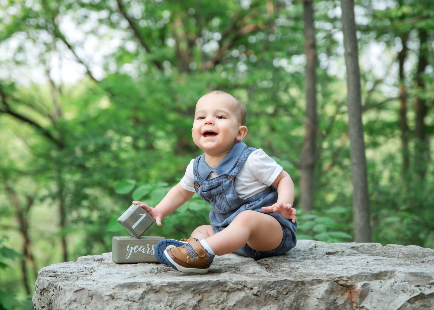 A young child in overalls sits on a large rock in the woods, smiling and laughing with joy in the green forest setting.