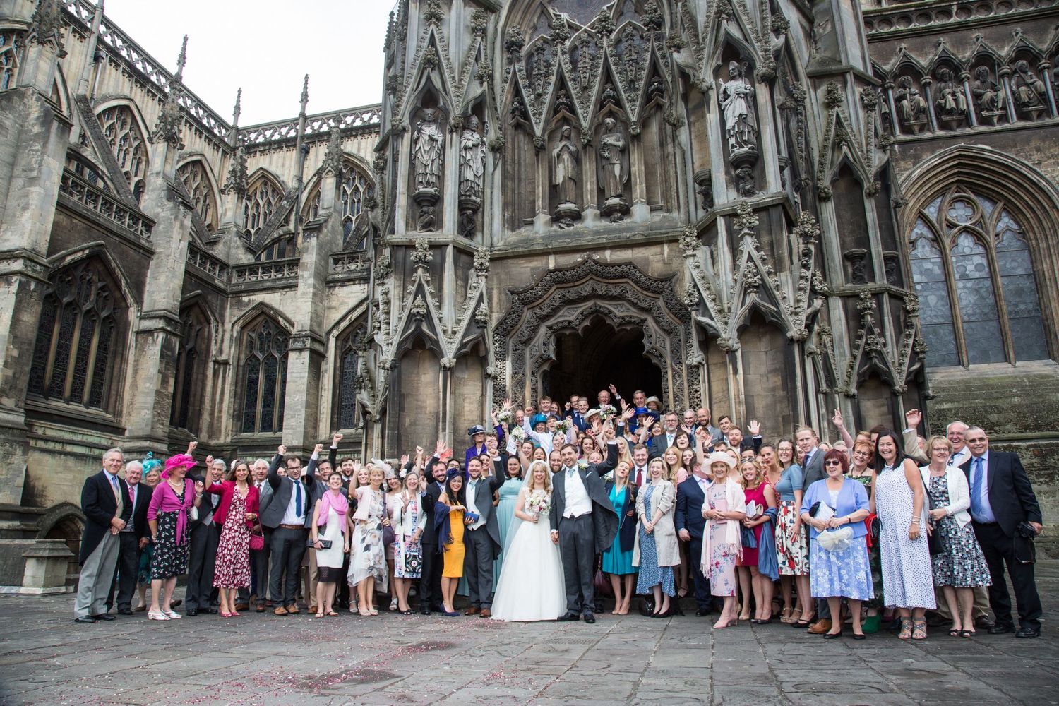 A wedding party gathers for group photos outside an ornate Gothic cathedral with intricate architectural details.