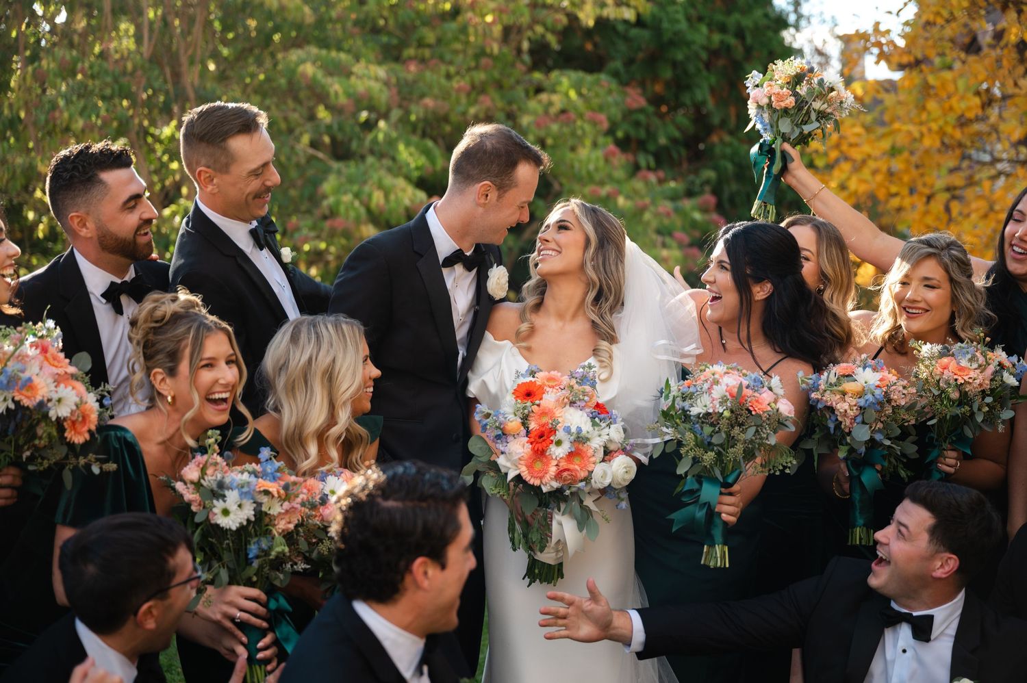 A wedding party celebrates outdoors in autumn with colorful fall foliage and floral bouquets. Captured by NYC wedding photographer.