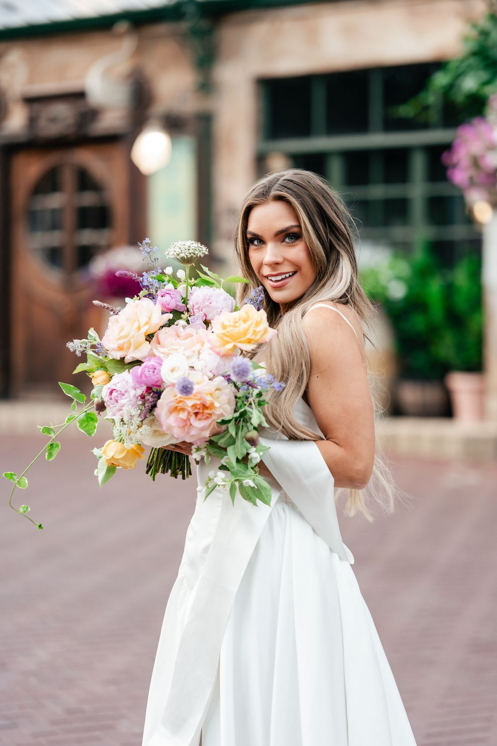A person in a white wedding dress holds a vibrant bouquet of peach, pink and purple flowers on a brick pathway.