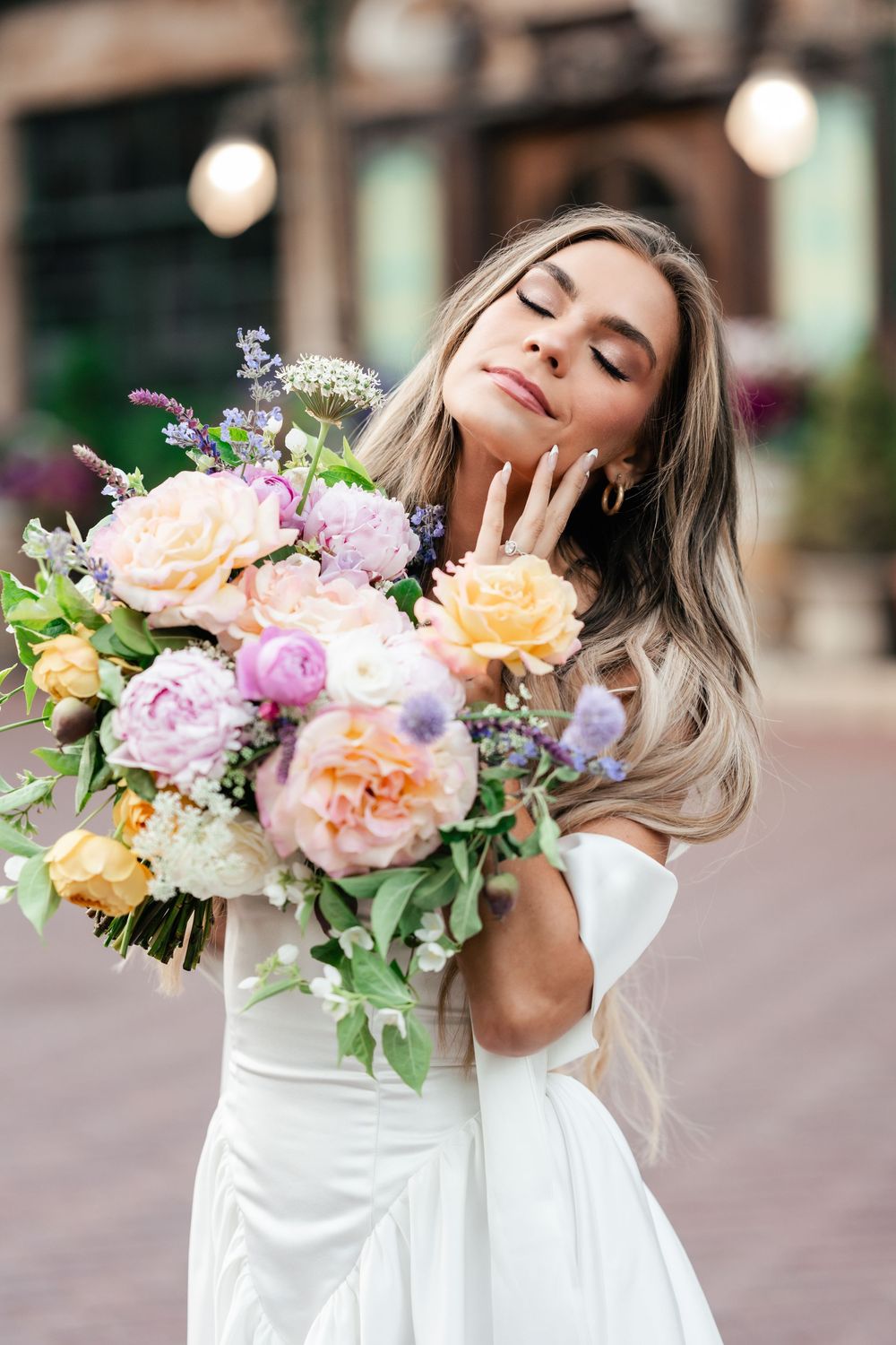 Person in white dress holds luxurious bouquet of peach, pink and purple flowers with eyes closed outside.