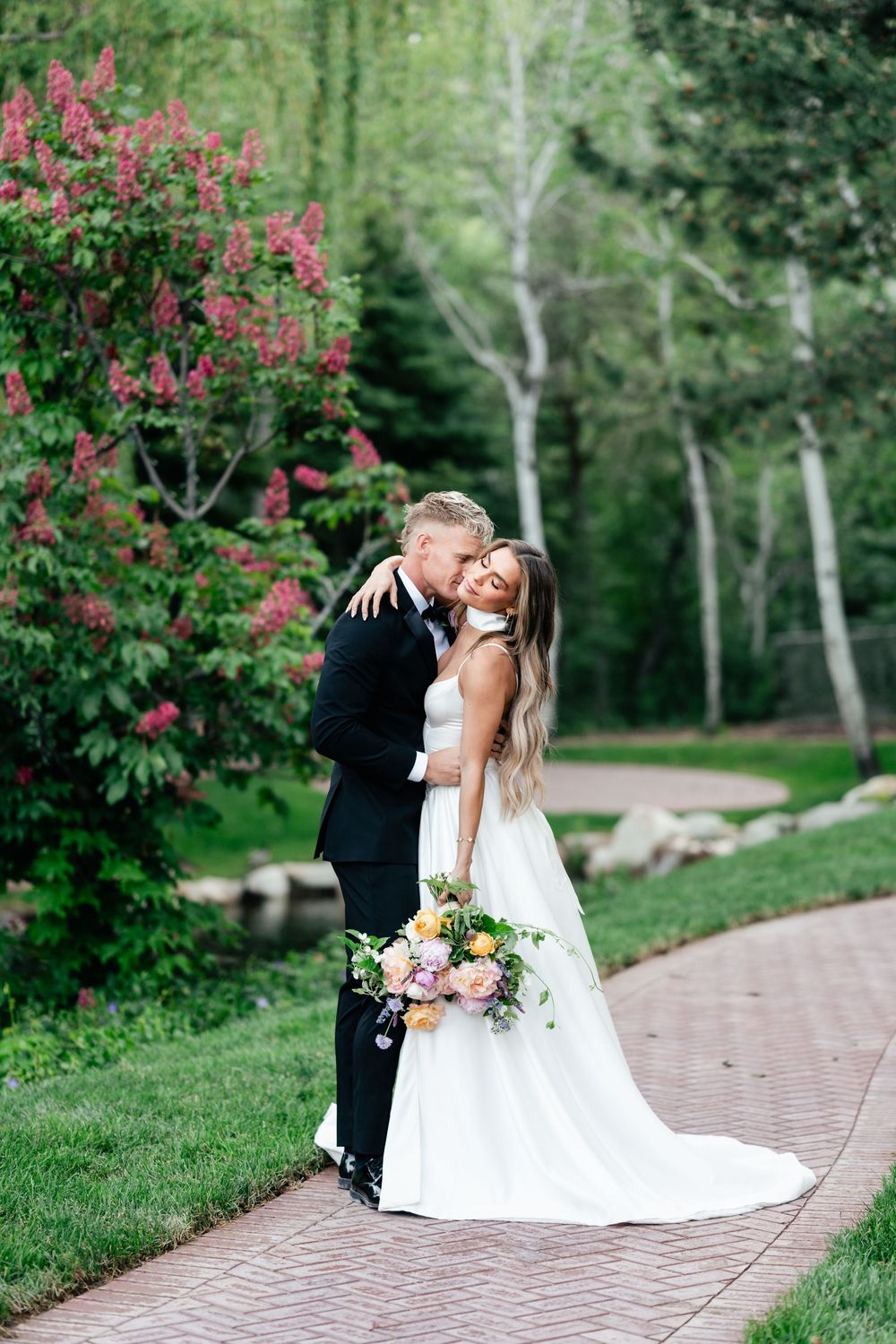 Newlyweds share romantic kisses under blooming pink garden flowers on a winding brick pathway.