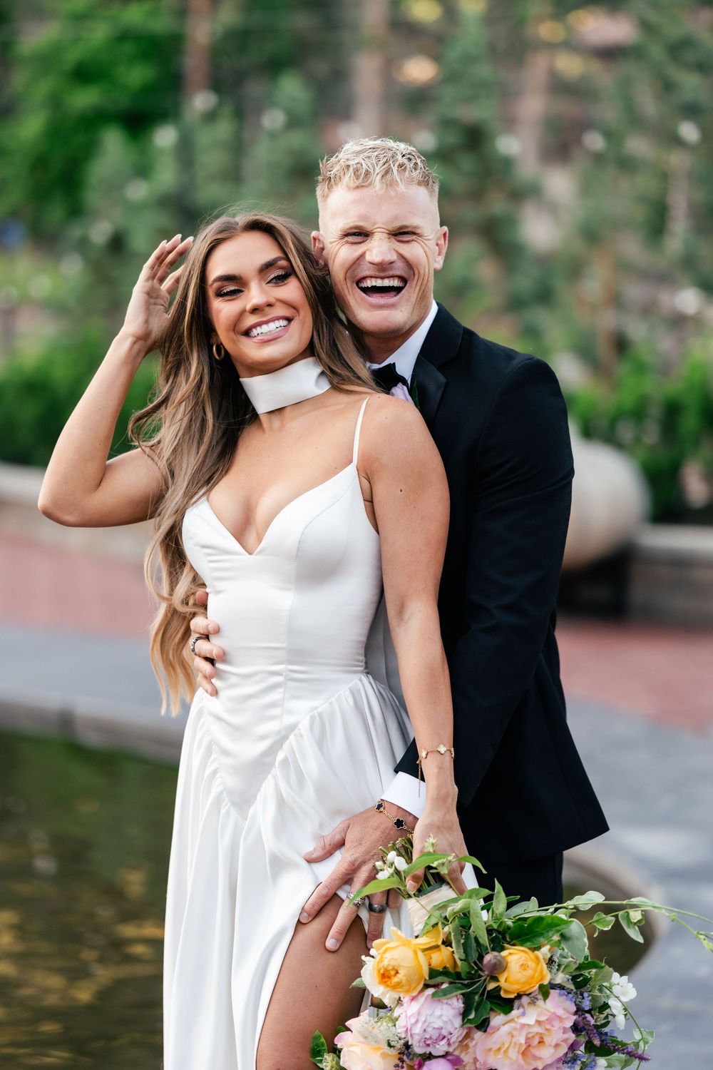 Happy couple sharing romantic moments outdoors with bride in white dress holding colorful bouquet of roses and flowers.