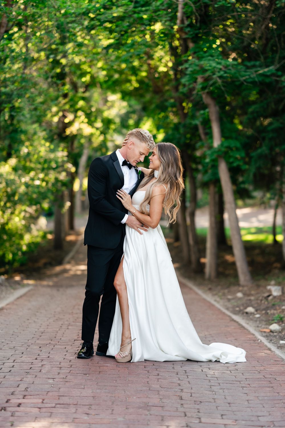 Bride and groom share romantic kisses on brick pathway surrounded by lush green trees in outdoor wedding setting.