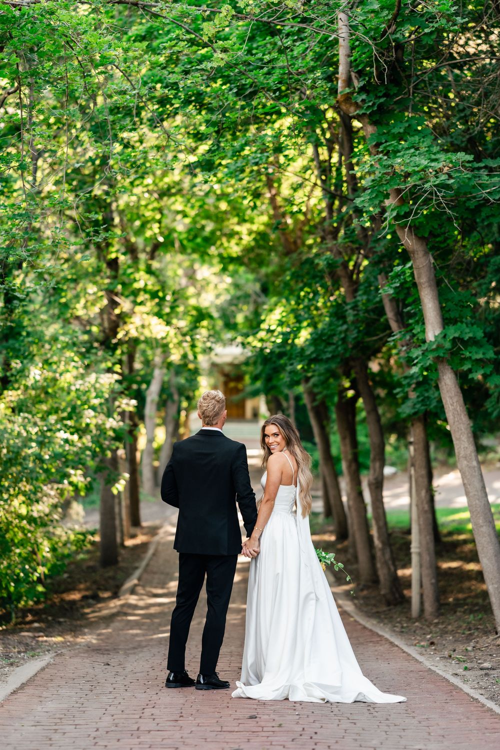 A couple walks along a tree-lined brick path in an elegant outdoor setting with dappled sunlight.