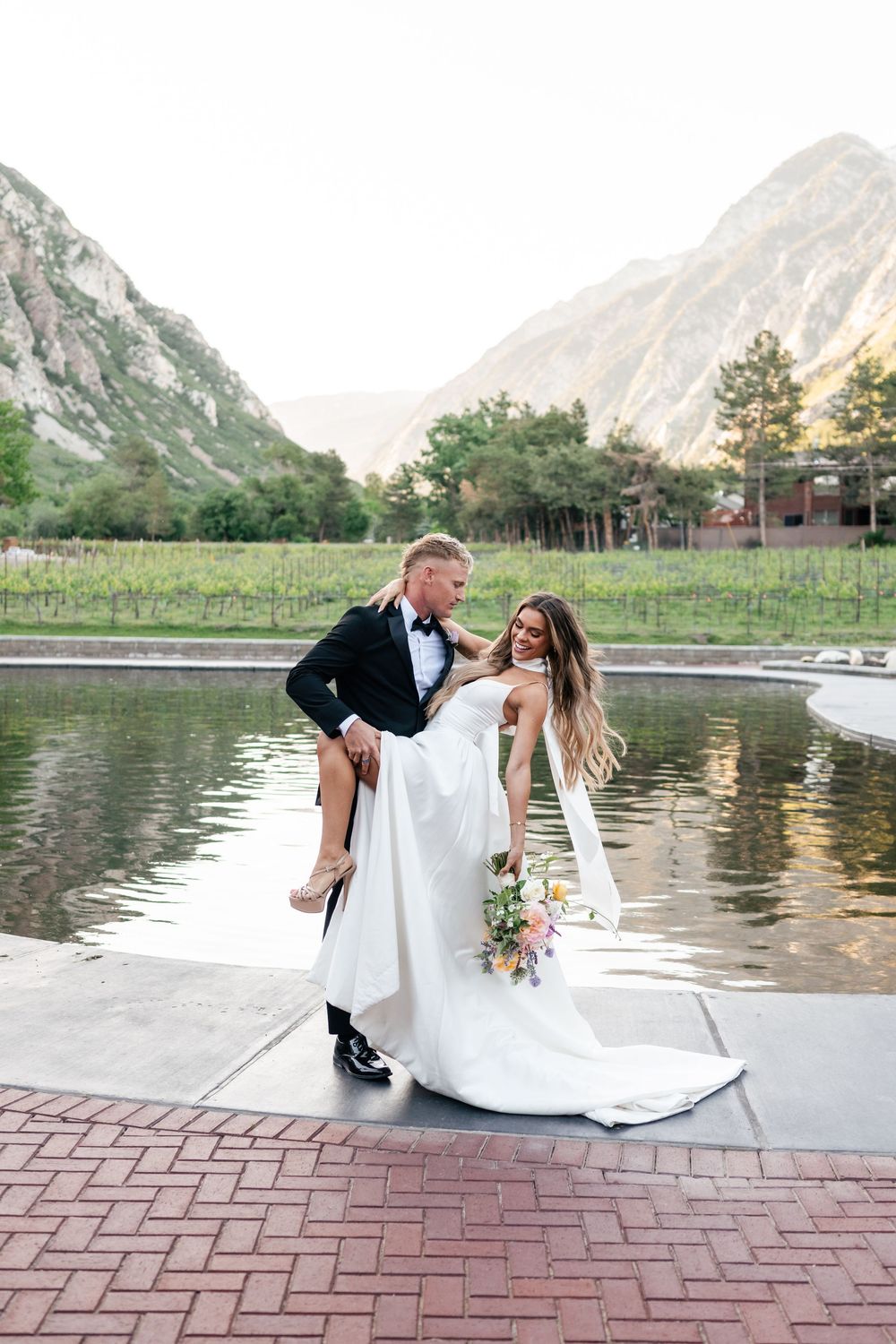 Newlyweds share a playful moment by a scenic mountain lake with the bride being carried across brick pavement.
