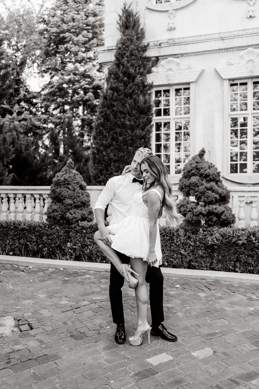 A couple in formal attire dances gracefully together on an outdoor stone patio near a white building with large windows.