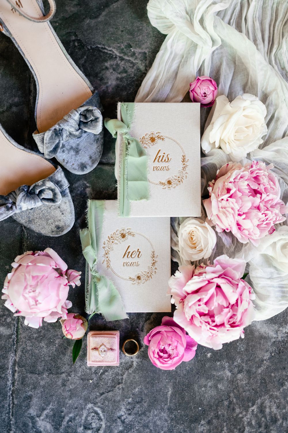 Wedding accessories with pink peonies, white roses, blush shoes and invitation cards with gold text on dark marble surface.