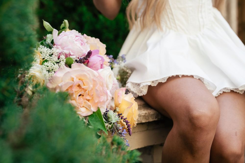A delicate bouquet of peach and pink roses with white flowers rests against a white dress in a garden setting.