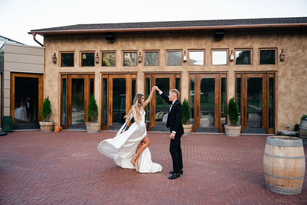 A couple dancing on a brick patio outside a rustic venue with glass windows and wooden barrels as decor.