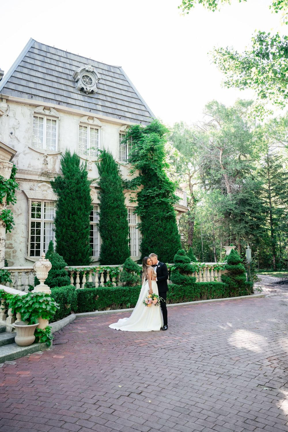 Elegant wedding couple stands on brick driveway in front of grand mansion with tall cypress trees and stone balustrade.