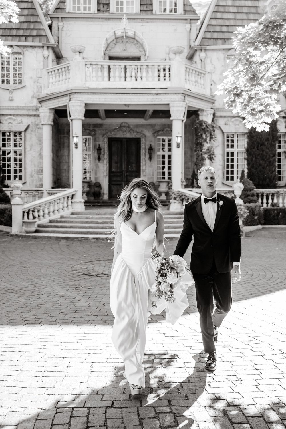 Black and white photo of wedding couple exiting luxurious mansion with grand pillared entrance and ornate balcony.