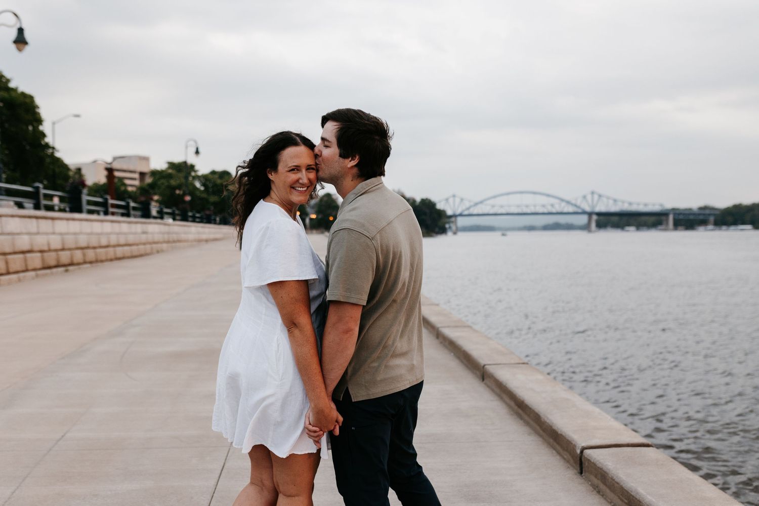 A newlywed couple walks along a lakeside promenade while sharing an intimate moment together at sunset.