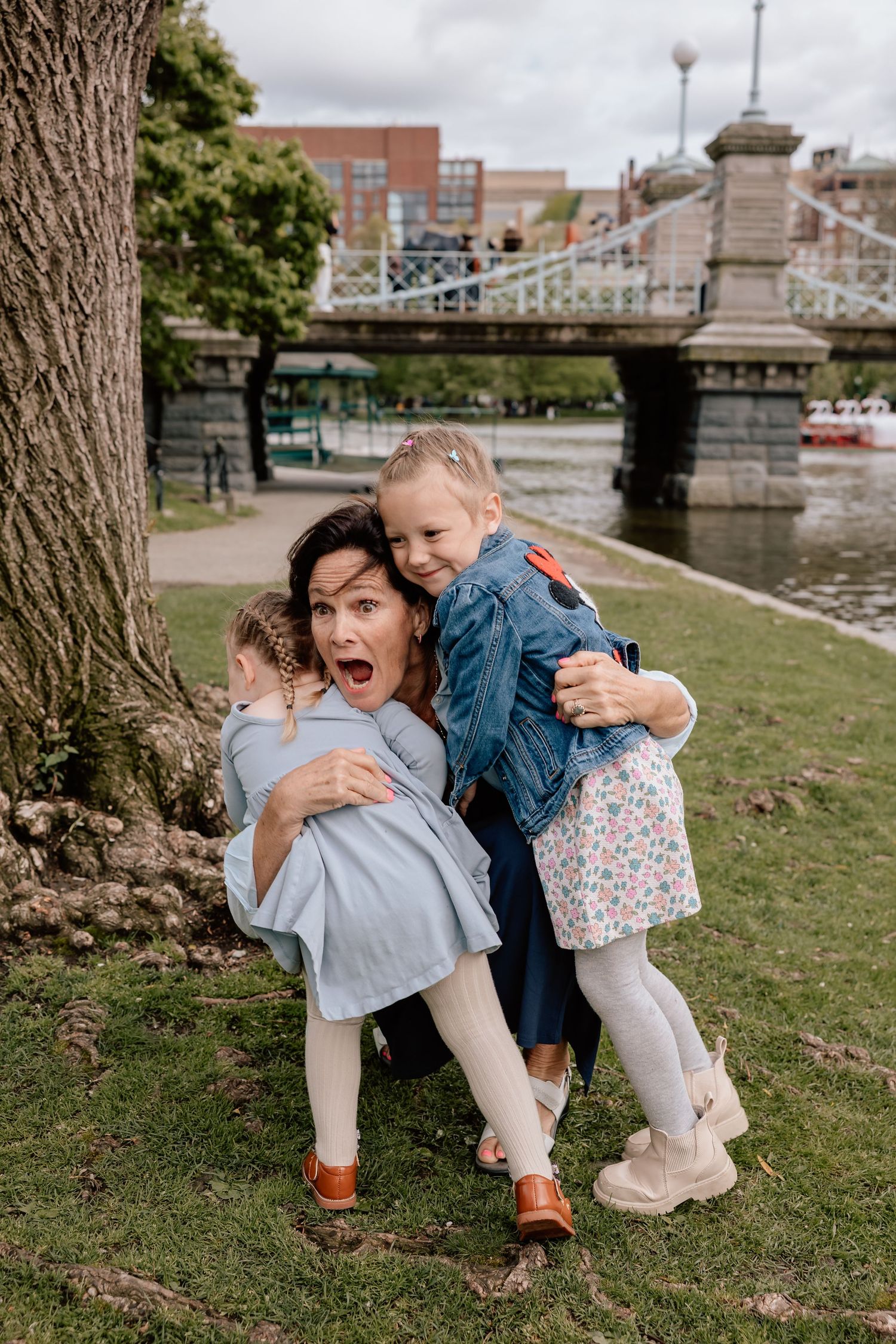Playful scene by a river with a historic bridge in the background as three people share a joyful moment on the grass.