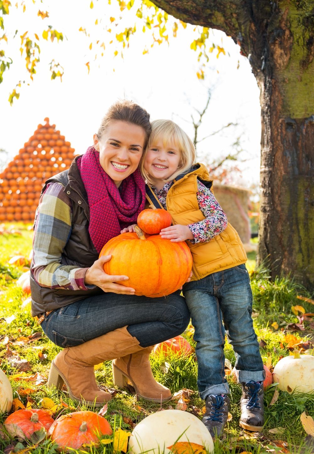 Two people in cozy autumn attire kneel in fallen leaves while holding large orange pumpkins in a sun-dappled outdoor setting.