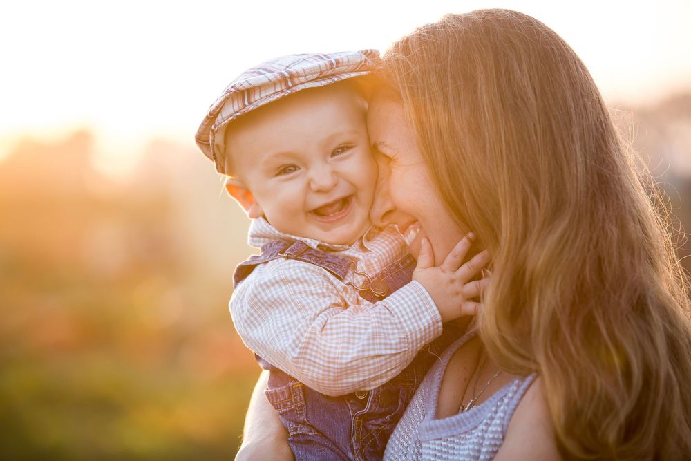 An adorable baby wearing a flat cap laughs joyfully with their mother during a warm golden sunset outdoors.