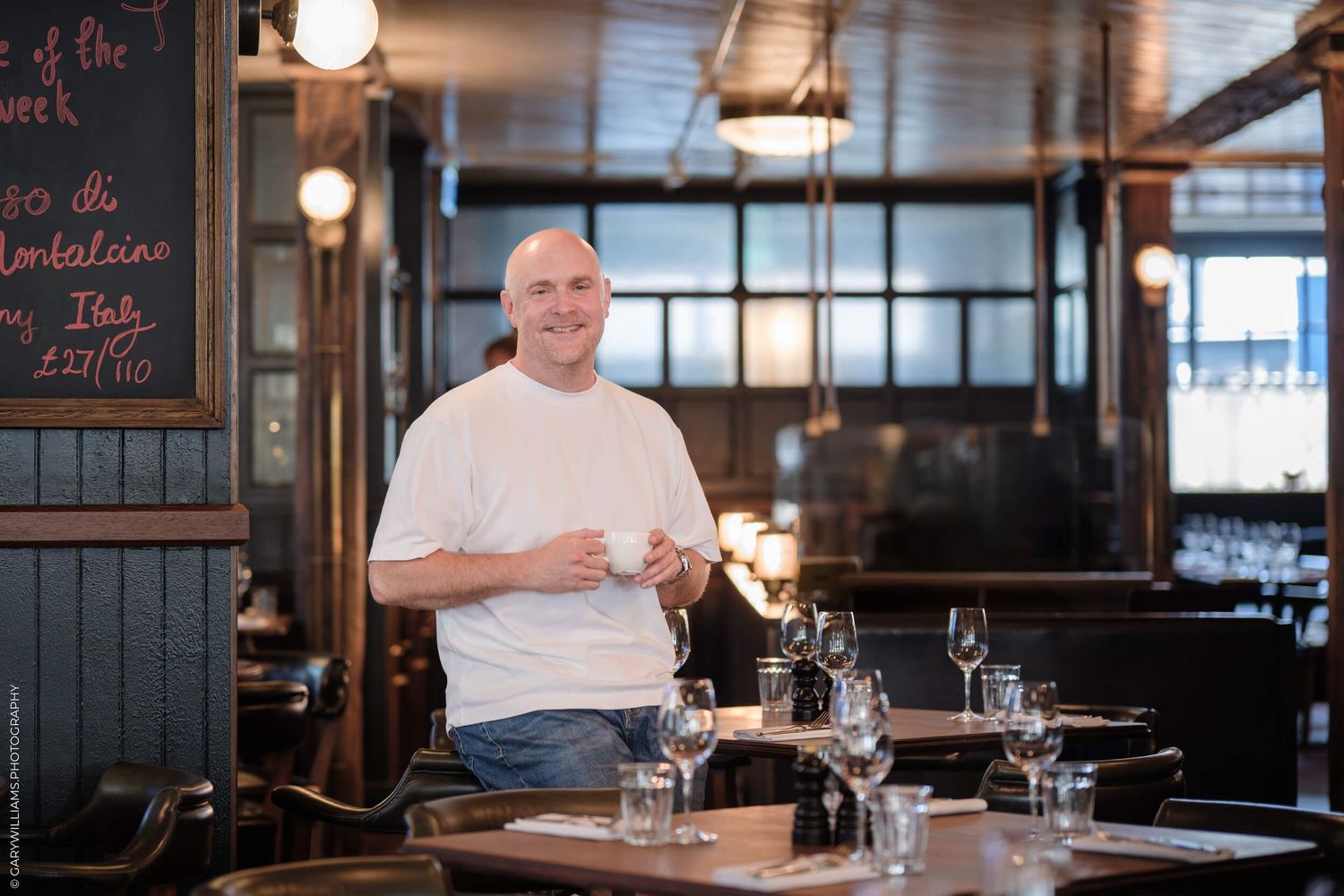 A chef in white attire leans casually on a counter in a restaurant dining room with warm lighting and table settings.