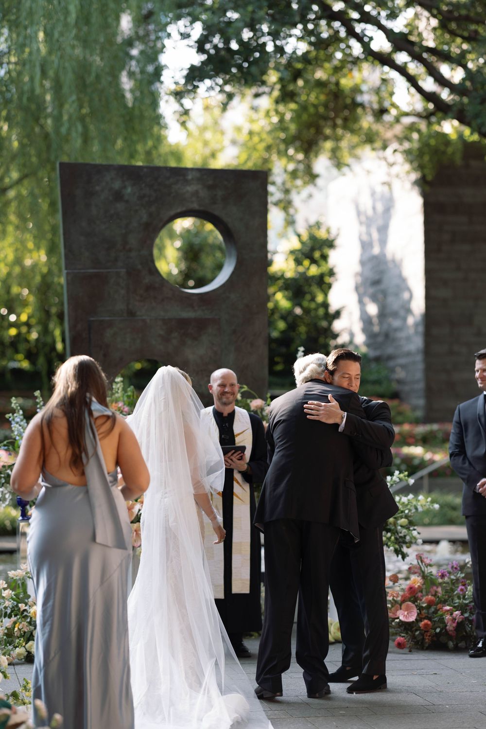 An outdoor wedding ceremony takes place in a garden setting with a modern stone sculpture featuring a circular opening.