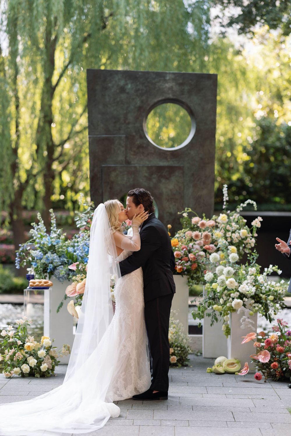Romantic outdoor wedding ceremony with couple kissing under modern stone arch surrounded by lush floral arrangements.