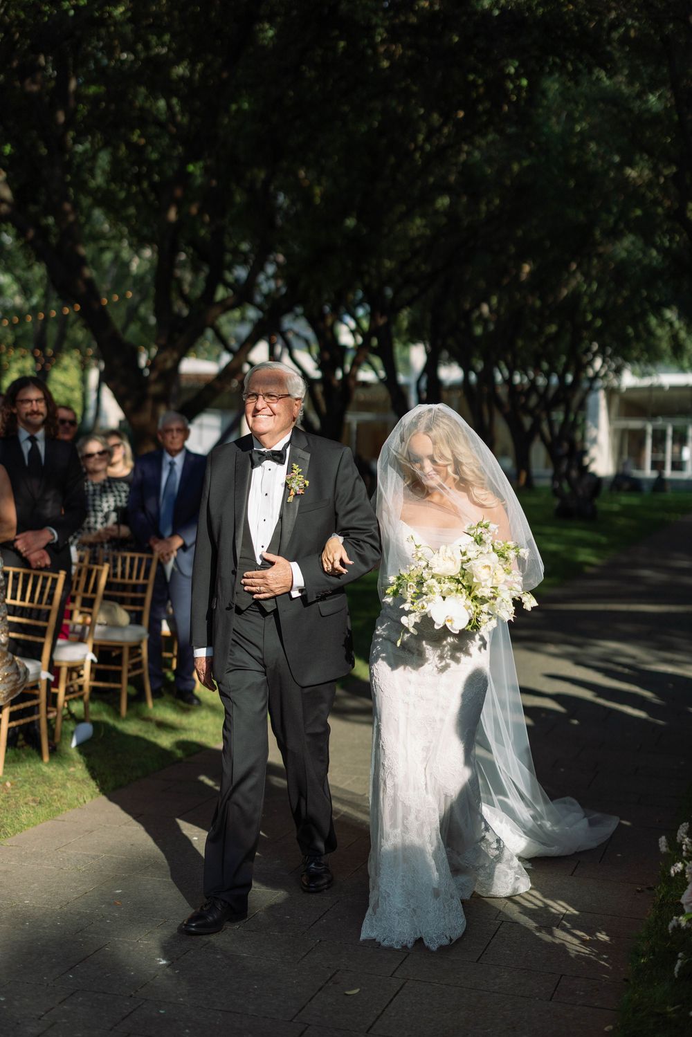 A bride and groom walk together on a tree-lined path during an outdoor wedding ceremony.