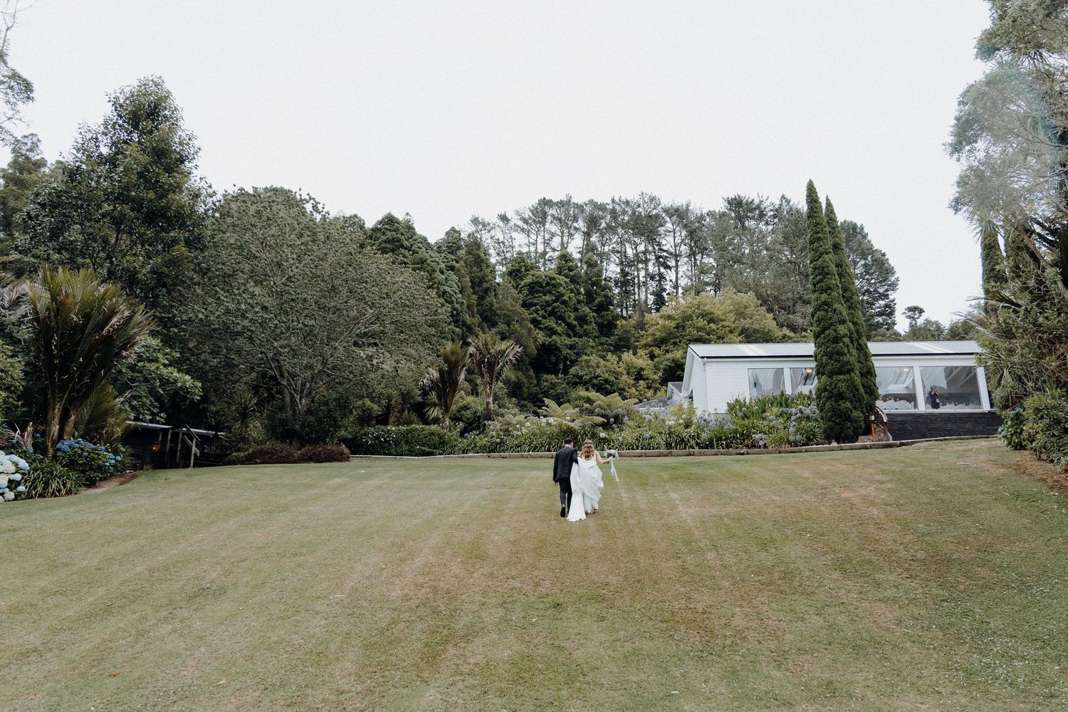 A couple strolls on a vast green lawn surrounded by cypress trees and lush gardens captured by Apertured Media Auckland Wedding Photographer