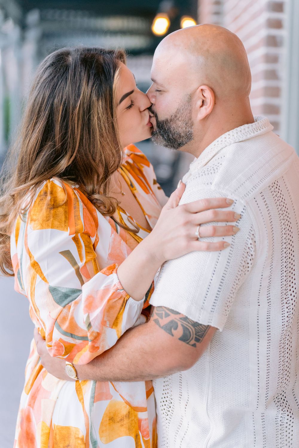A couple sharing a romantic kiss outdoors, wearing a yellow patterned dress and white knit sweater.
