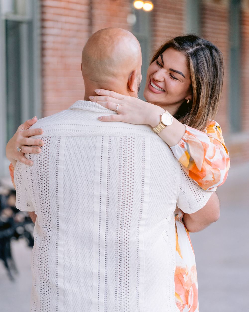 Two people share a warm embrace while smiling and wearing light-colored clothing on a sidewalk near a brick building.