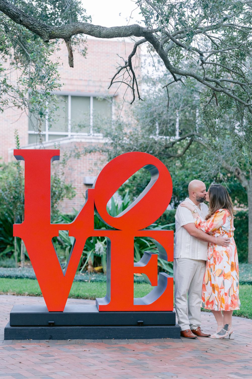 A couple stands beside a large red LOVE sculpture in a garden setting with overhanging tree branches.