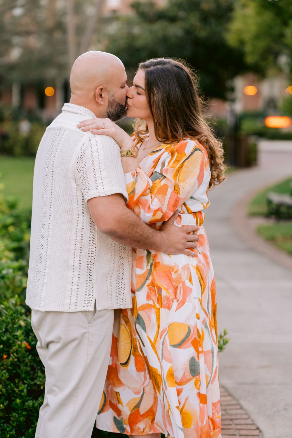A couple shares a romantic kiss on a garden path at dusk, with the woman wearing a vibrant orange floral dress.