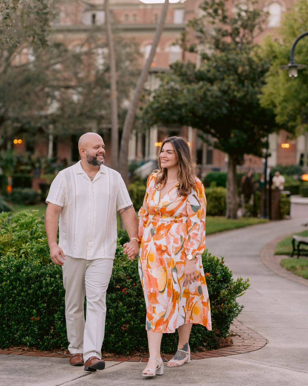 A couple strolls along a winding garden path, wearing light summer attire with brick buildings and trees in the background.