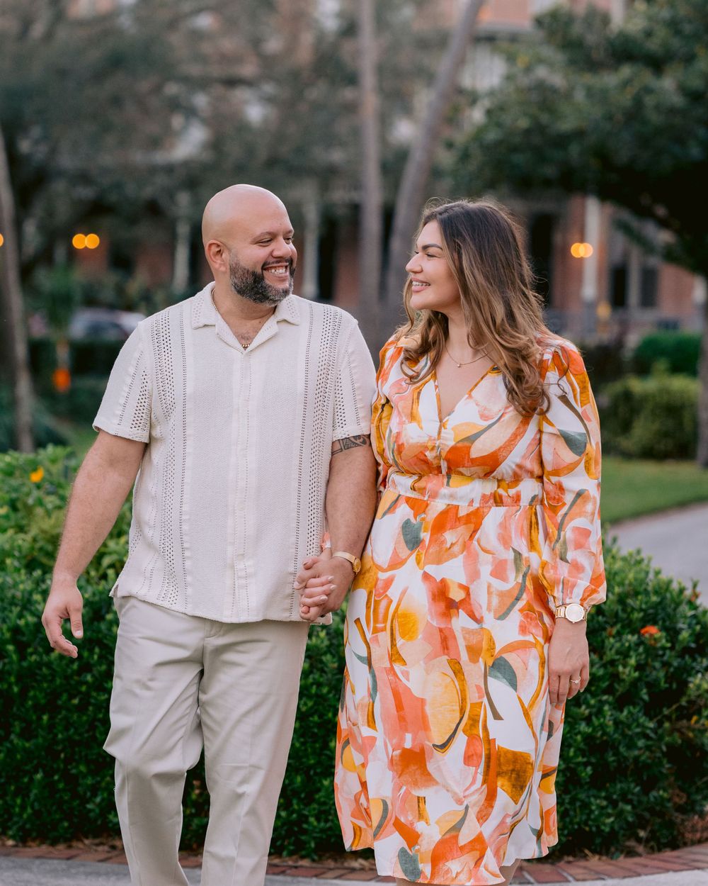 A couple walks on a garden path at dusk, with her wearing a floral orange dress and him in light colored clothing.
