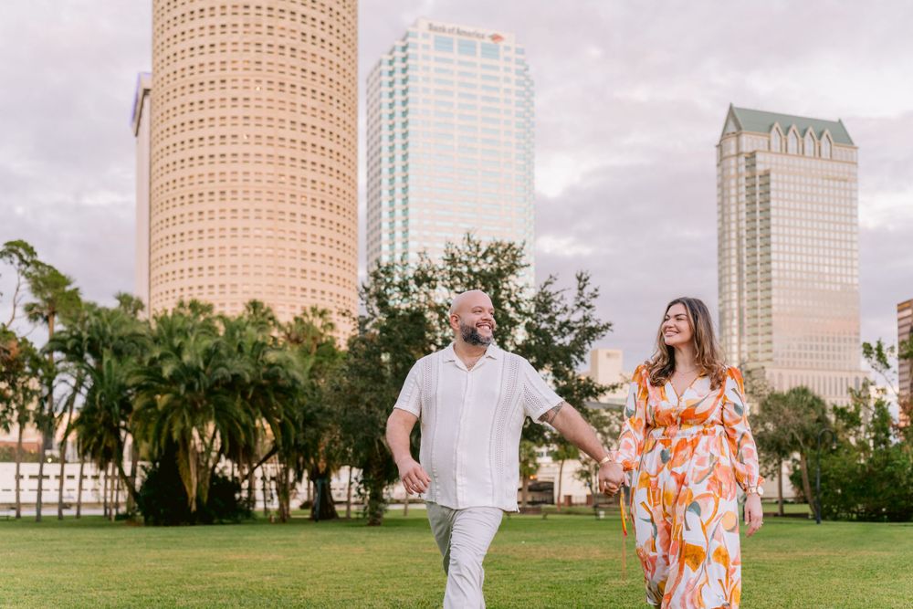 Happy couple walks together on a green lawn with downtown Tampa high-rise buildings in the background.