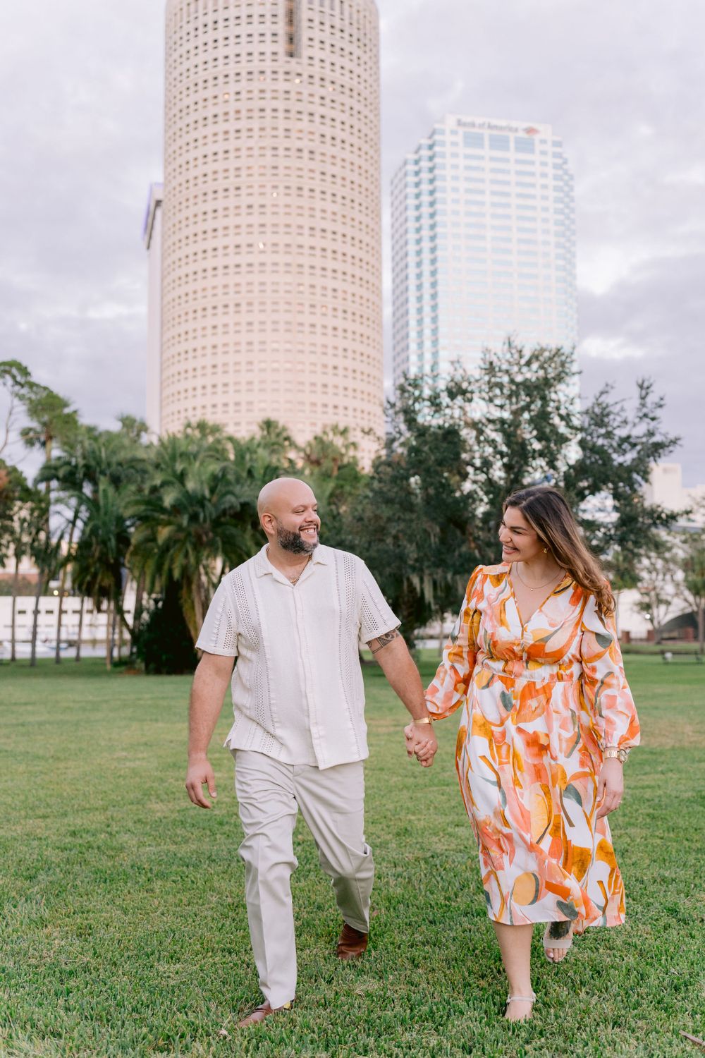 A couple walks hand in hand through a grassy park with downtown skyscrapers and palm trees in the background.