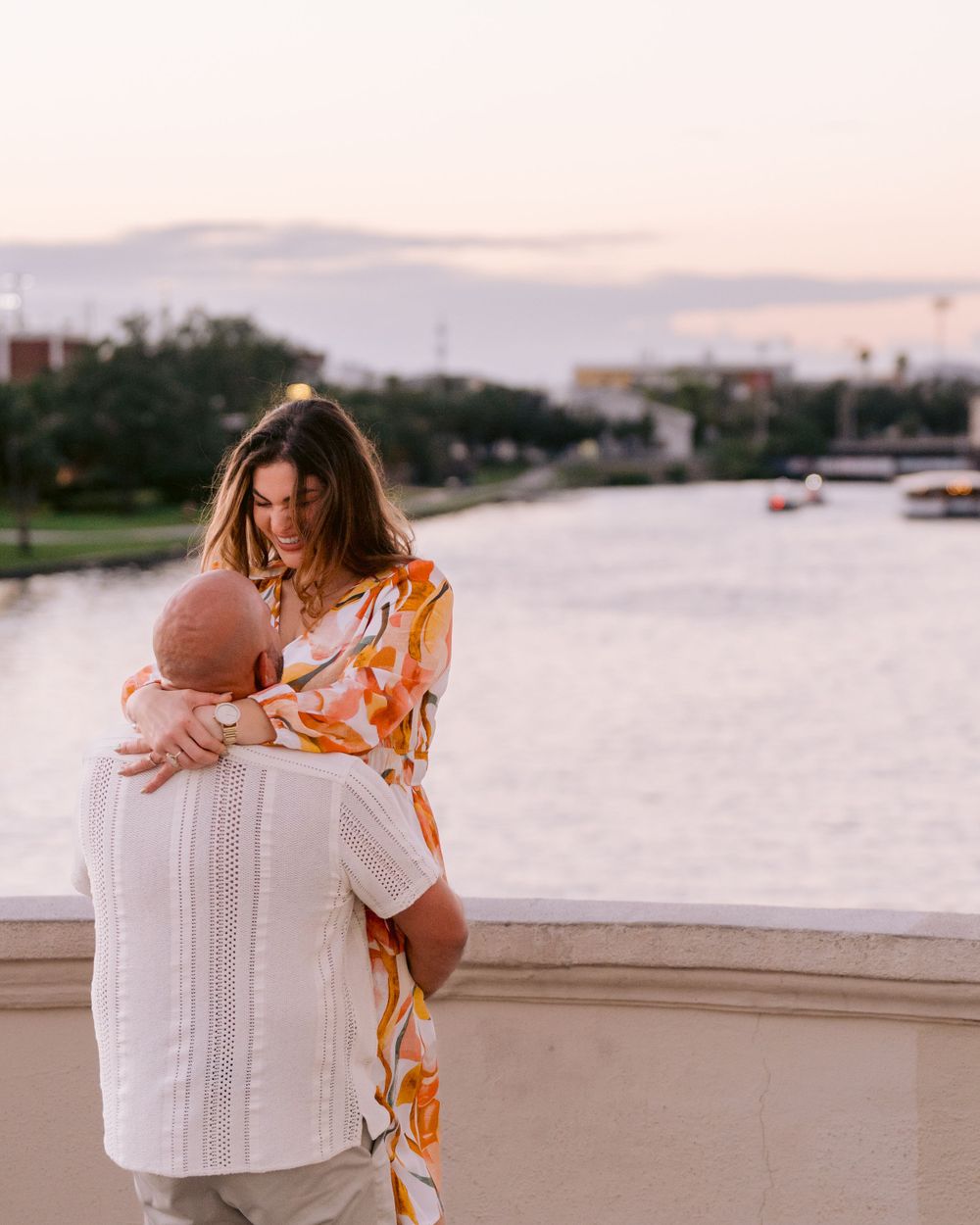 A couple embraces on a waterfront balcony at sunset, with boats visible on the calm water below.