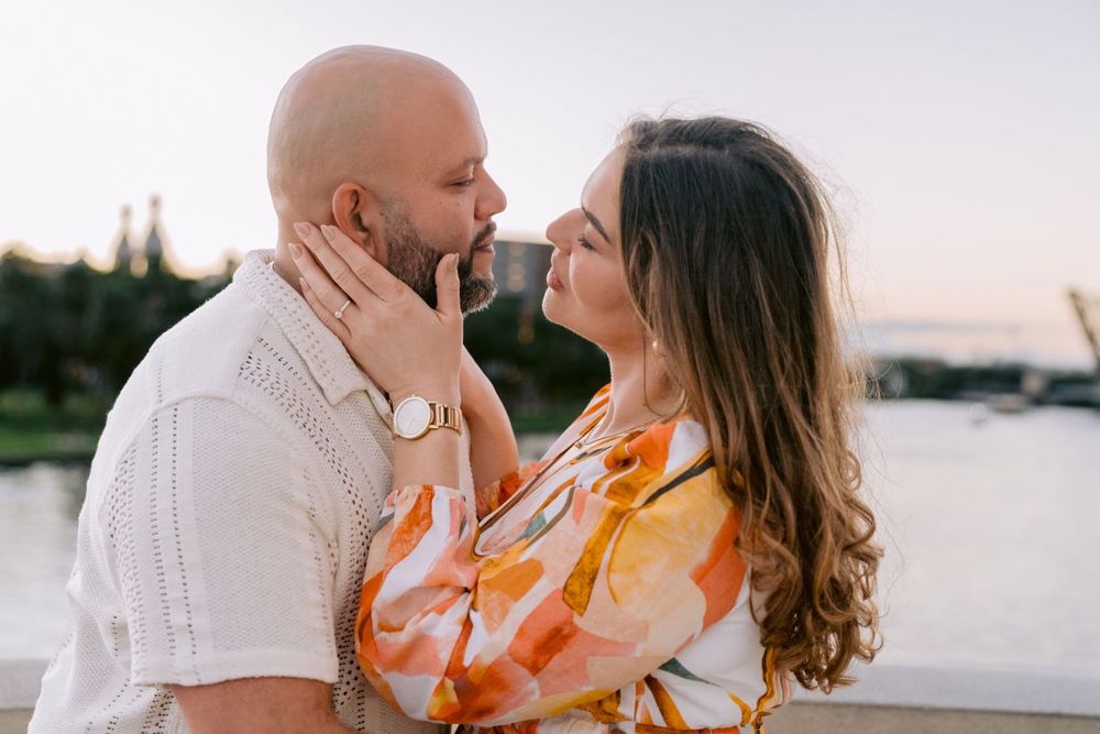 Couple in romantic embrace by waterfront at sunset, with woman in orange floral dress and man in white shirt.